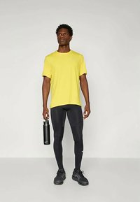 Mees kollases spordisärgis ja mustades retuusides hoiab musta veepudelit, seistes ühetaolise heleda tausta ees.