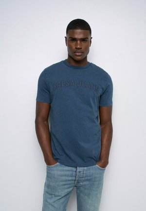 Hombre con pelo corto lleva una camiseta azul "Salsa Jeans" y vaqueros azul claro, de pie con las manos en los bolsillos contra un fondo blanco liso.