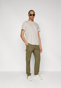 Striped short-sleeve t-shirt in light beige and brown, olive cargo pants with side pockets, beige sneakers, and green sunglasses.