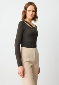 Maglietta a maniche lunghe, aderente, di tessuto marrone scuro con un dettaglio a taglio sullo scollo, abbinata a pantaloni beige su misura. Tessuto liscio.