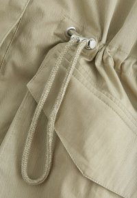 Tissu beige avec un cordon tressé enfilé à travers des œillets métalliques argentés au-dessus d'une poche avant avec des coutures visibles.