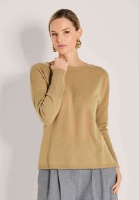 Frau in einem beigen Langarmpullover und grauen Hosen, mit großen goldenen Creolen, die mit einer Hand nahe ihrem Hals posiert.