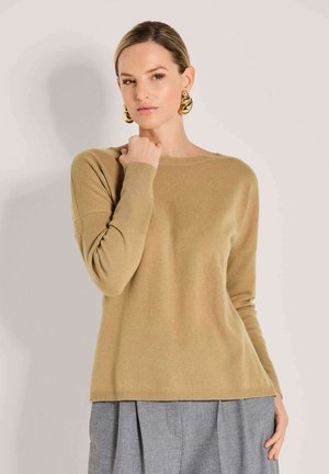 Frau in einem beigen Langarmpullover und grauen Hosen, mit großen goldenen Creolen, die mit einer Hand nahe ihrem Hals posiert.