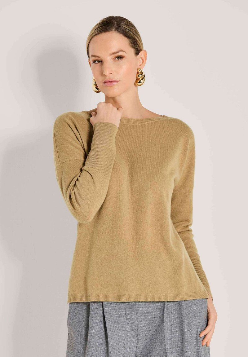 Frau in einem beigen Langarmpullover und grauen Hosen, mit großen goldenen Creolen, die mit einer Hand nahe ihrem Hals posiert.