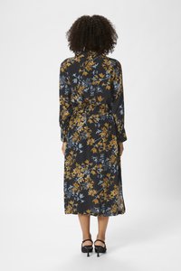 Floral dress in black fabric with blue and mustard patterns, long sleeves, belted waist, and side slits, paired with black heeled sandals.