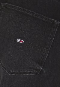 Un tissu en denim noir présente une poche cousue avec un petit logo rectangulaire de Tommy Hilfiger en rouge, blanc et bleu.