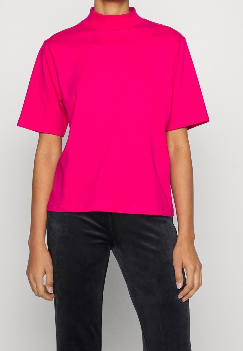 Personne portant un haut rose vif à manches courtes et col montant factice, ainsi qu’un pantalon noir, debout devant un fond clair uni.