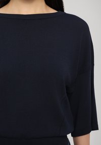 Femme portant un top en maille bleu marine foncé à manches courtes avec un large col rond, montrée du cou à la taille.