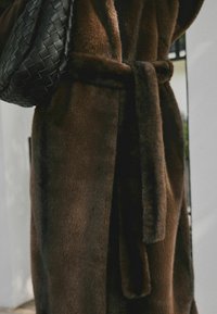 Brown faux fur robe with a matching belt and a textured black woven bag slung over one shoulder, against a neutral backdrop.