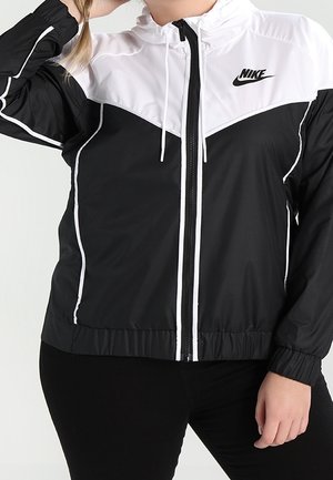 Kobieta w czarno-białej wiatroszczelnej kurtce Nike na zamek z białymi lamówkami i elastycznymi mankietami, pozująca z jedną ręką przy głowie.