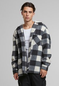 Camicia in flanella a quadri testurizzati nei colori nero, grigio e bianco; presenta un cappuccio, chiusura con bottoni e una tasca frontale sul lato sinistro.