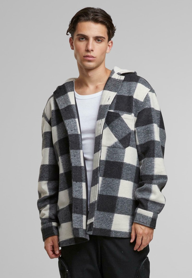 Camicia in flanella a quadri testurizzati nei colori nero, grigio e bianco; presenta un cappuccio, chiusura con bottoni e una tasca frontale sul lato sinistro.
