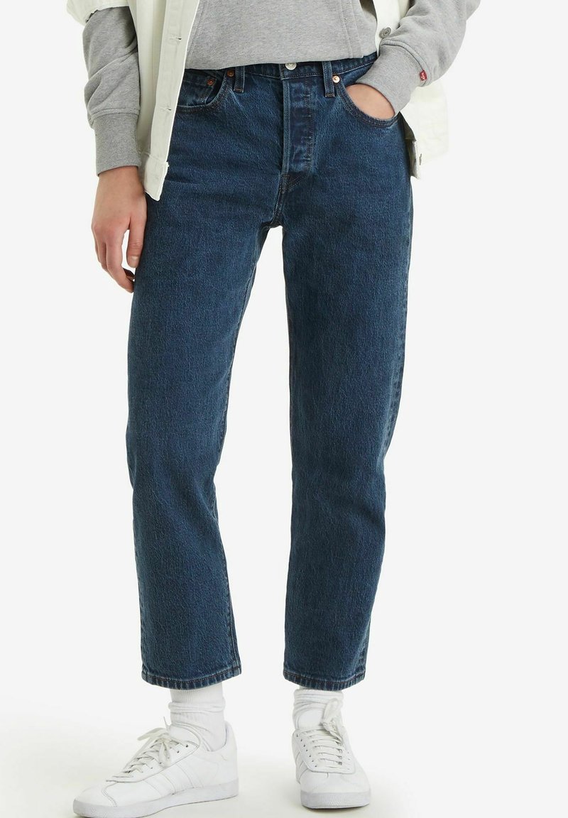Personne portant un jean bleu droit, un sweat-shirt gris, une veste blanche, des chaussettes blanches et des baskets blanches, main dans la poche sur fond uni.