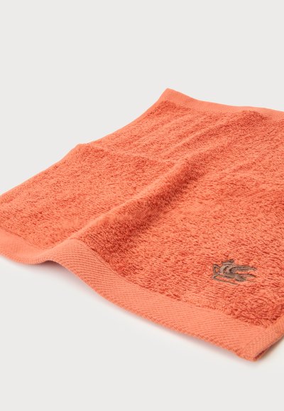 Serviette en coton texturé orange avec un petit logo gris brodé dans un coin sur une surface blanche.