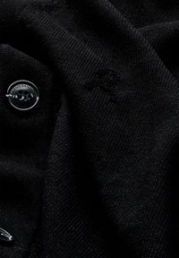 Schwarzer Strickstoff mit weicher Textur, ausgestattet mit einem runden schwarzen Knopf, der mit "ARMEDANGELS" geprägt ist, sowie einem dezenten Logodetail.