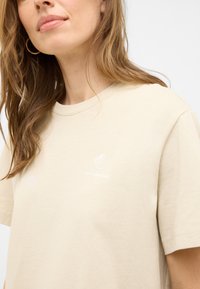 Beige cotton T-shirt with a crew neck and short sleeves. Features a white shell graphic and text "en mer." Smooth texture.