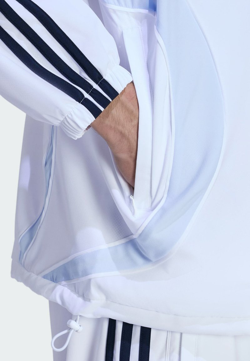 Veste de sport blanche avec des rayures bleu marine foncé. Comprend une poche latérale zippée et un cordon ajustable à l'ourlet. Texture de tissu lisse.