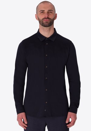 Uomo con barba corta che indossa una camicia nera a maniche lunghe con bottoni e pantaloni scuri con motivo su uno sfondo chiaro e uniforme.
