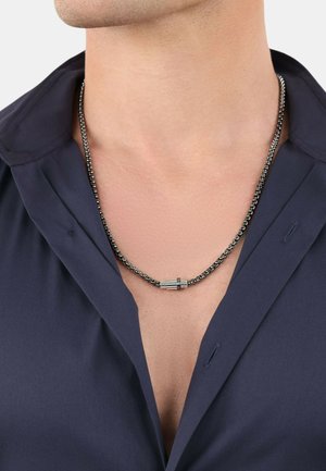 Collana in metallo con catena nera e accenti cilindrici in argento. La catena ha una finitura testurizzata e poggia su una camicia di tessuto scuro.