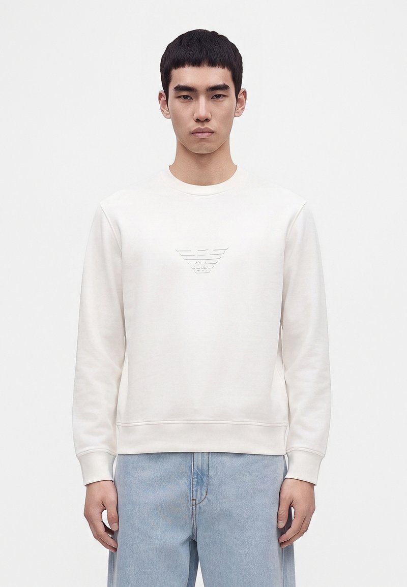 Jeune homme portant un sweat-shirt blanc avec logo en relief sur la poitrine et un jean bleu clair, debout devant un fond blanc uni.