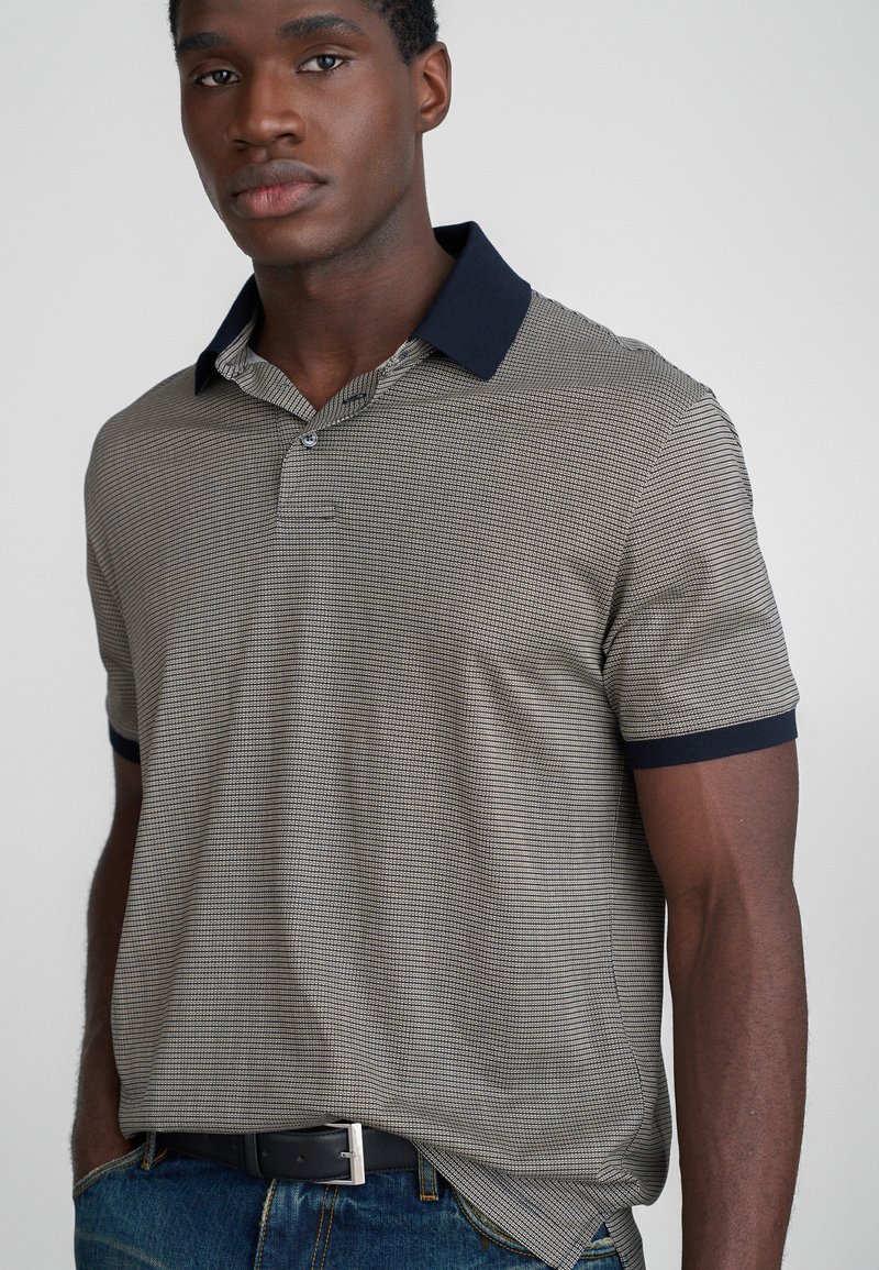 Vyriška trumpo rankovio polo marškiniai su raštuotu pilku ir tamsiai mėlynu dizainu, vientisa tamsiai mėlyna apykakle ir tekstūruotu audiniu. Laisvas kirpimas, derinami su mėlynais džinsais.