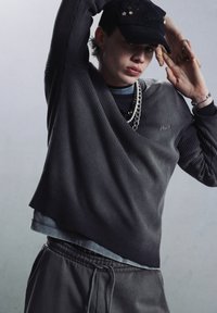 Donker geribbeld langwerpig shirt, gelaagd over een grijs T-shirt, gecombineerd met grijze joggingsbroek. Accessoires omvatten een zwarte pet met versieringen en kettingen.