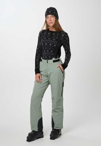 Vrouw met een zwart patroon langemouwen top, lichtgroene sneeuwbroek met bretels, zwarte skischoenen en een bijpassende zwarte patroon muts.