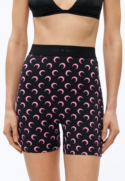 Donna che indossa pantaloncini neri a vita alta con motivo di lune crescenti rosa e un top corto nero su sfondo bianco.