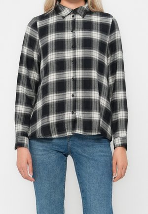 Femme portant une chemise à carreaux noire et blanche à manches longues boutonnée et un jean bleu, debout devant un fond uni.