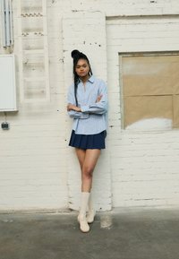 Lyseblå stribet skjorte med knapper, marineblå plisseret nederdel, cremede sokker og beige sneakers. Model poserer foran en hvid murstensvæg.