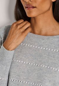 Frau mit mittelbrauner Haut und langen dunklen Haaren trägt einen hellgrauen Strickpullover mit horizontalen offenen Maschen am Halsauschnitt und an den Ärmeln.