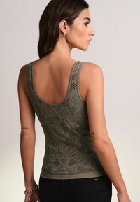 Olijfgroene tanktop met textuur en bloemmotief, slim fit ontwerp, brede schouderbanden en geribbelde zoom. Draagt met zwarte broek.