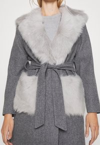 Woman wearing a gray belted coat with light gray faux fur collar and large faux fur pockets, standing against a white background.