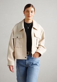 Beige zkrácená bunda s velkými předními kapsami, špičatým límcem a zipem. Nosit se přes černý top, v kombinaci se světlými modrými džíny.