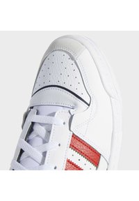 Gros plan d'une basket blanche en cuir avec des bandes rouges, une boîte à orteils perforée, des lacets blancs et un détail en daim sur le bout.
