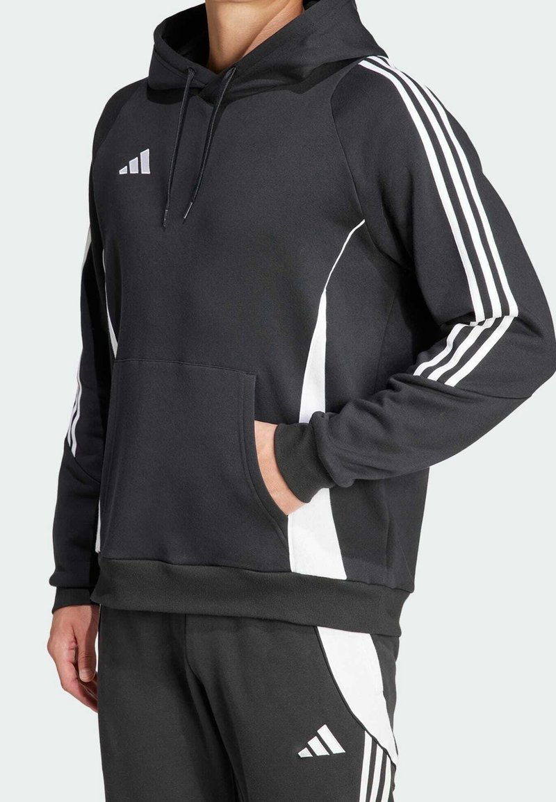 Personne portant un sweat à capuche noir et un pantalon de jogging avec des rayures et logos blancs, main dans la poche du sweat, sur fond uni.