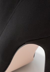 Gros plan d'une chaussure à talon haut en tissu noir texturé montrant le talon et les détails de la semelle incurvée.