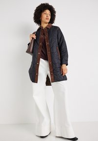 Kvadrokrpna črna jakna z rjavimi kordnimi detajli in gumbi na pritisak, kombinirana s temnim zgornjim delom in belimi širokimi hlačami. Gladka tekstura, rokavi do rame.