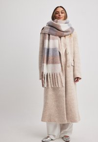 Beige villakangas takki, jossa on pehmeä rakenne, kaksirivinen malli ja suuri kaulus. Harmaa ja ruskea kuvioitu huivi, jossa on fringed yksityiskohtia.