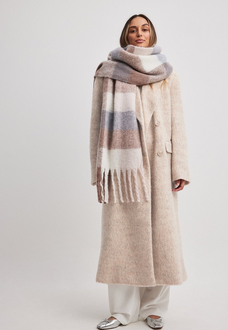 Beige villakangas takki, jossa on pehmeä rakenne, kaksirivinen malli ja suuri kaulus. Harmaa ja ruskea kuvioitu huivi, jossa on fringed yksityiskohtia.