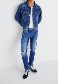 Person wearing blue denim jacket, distressed blue jeans, white t-shirt, and black high-top sneakers with white laces standing against white wall.