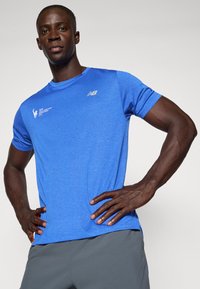 Zila sporta t_krekls ar īsām piedurknēm, teksturēta auduma, atstarojošs New Balance logo un Ņujorkas maratona zīmols. Apvienots ar pelēkiem šortiem.