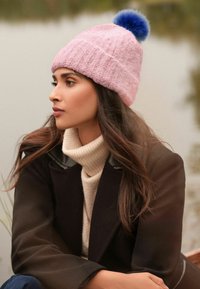 Pehmea roosa kootud müts sinise faux karusnahast pom-pom-iga. Mudel kannab beeži kõrge kaelusega sviitšerit ja musta jope, rõhutades mütsi tekstuuri.