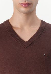 Hnedý sveter s výstrihom do „V“ z jemnej pleteniny, s malým logom v červenej, bielej a modrej farbe na hrudi. Hladká textúra a priliehavý strih.
