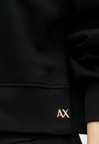 Μαύρος φούτερ με μεντεσέδες και τελείωμα, διακοσμημένος με μικρή χρυσή λεπτομέρεια "AX" κοντά στον καρπό στην δεξιά πλευρά.