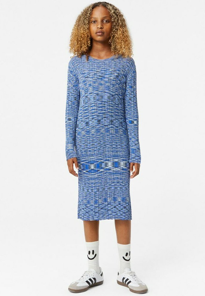 Sininen neulemekko, jossa on pitkät hihat ja teksturoitu kuvio. Yhdistetty valkoisiin sukkiin ja ruskeisiin/valkoisiin tennareihin.