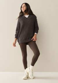 Tumehall suurte mõõtmetega kootud kampsun V-kaelusega, tekstuurse kangaga, soonikkoes ääreline, pruunide retuuside ja paksude valgete tossudega.