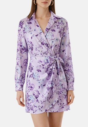 Femme portant une robe portefeuille lilas à manches longues avec un imprimé floral violet et un nœud noué à la taille, accessoirisée de boucles d'oreilles créoles en or.