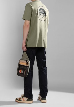 Oliivinvihreä puuvillainen t-paita, jossa vuoristokuvio selässä, yhdistettynä tummiin cargo-housuihin, kermaan ja vihreisiin lenkkareihin sekä pieneen harmaaseen olkalaukkuun.