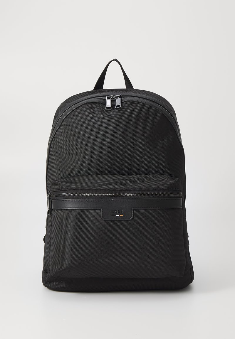 Μαύρο σακίδιο με επιφάνεια από υφαντό ύφασμα, καμπύλη κορυφή, δίδυκους ασημένιους φερμουάρ και μπροστινή τσέπη με φερμουάρ που διαθέτει πλακέτα με το λογότυπο.